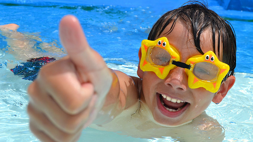 Ein lachender Junge mit Taucherbrille hebt im Schwimmbad seinen Daumen in die Luft.